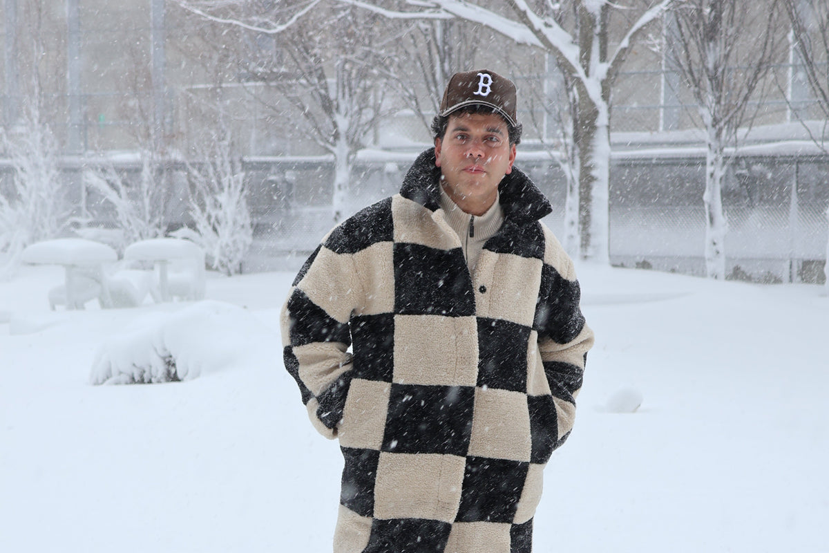 Miles Perry standing in deep snow wearing a long black and beige checkered coat, black pants, and a dark "Boston" baseball cap with a white 'B' logo, with snow-covered trees and a brick building in the background.